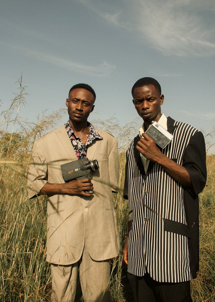 Two fashionable young men with vintage cameras pose confidently in a field, setting a retro yet modern vibe.