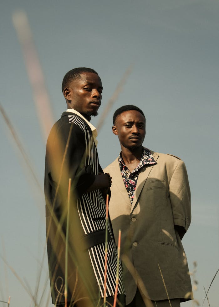 Two men in fashionable attire stand in a natural setting under a clear sky.