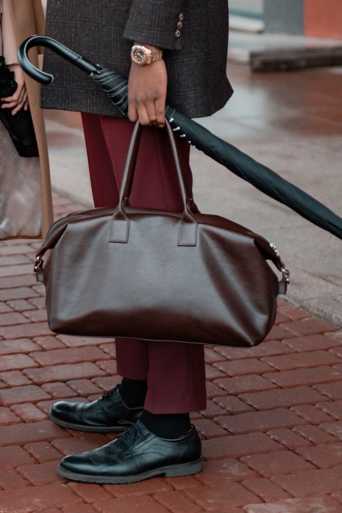 Stylish man holding an umbrella and a leather bag on a paved walkway, showcasing urban fashion.